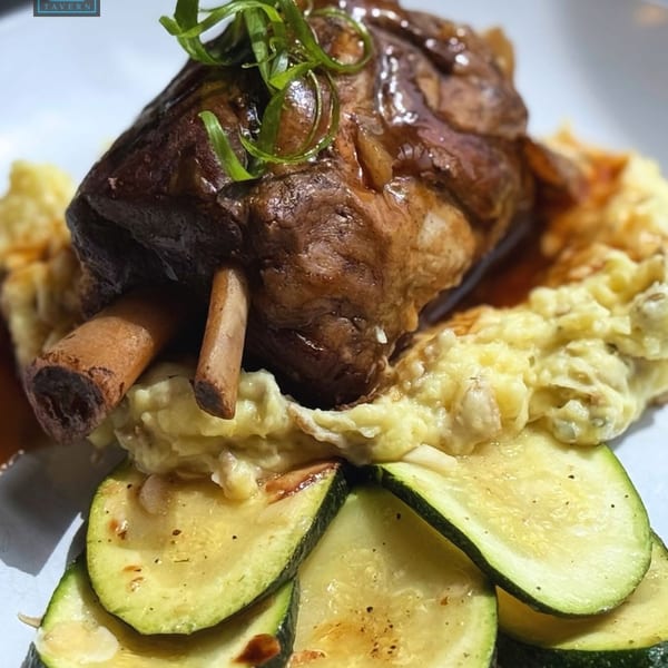 Braised lamb shank with mashed potatoes, gravy, and sliced zucchini on a white plate.