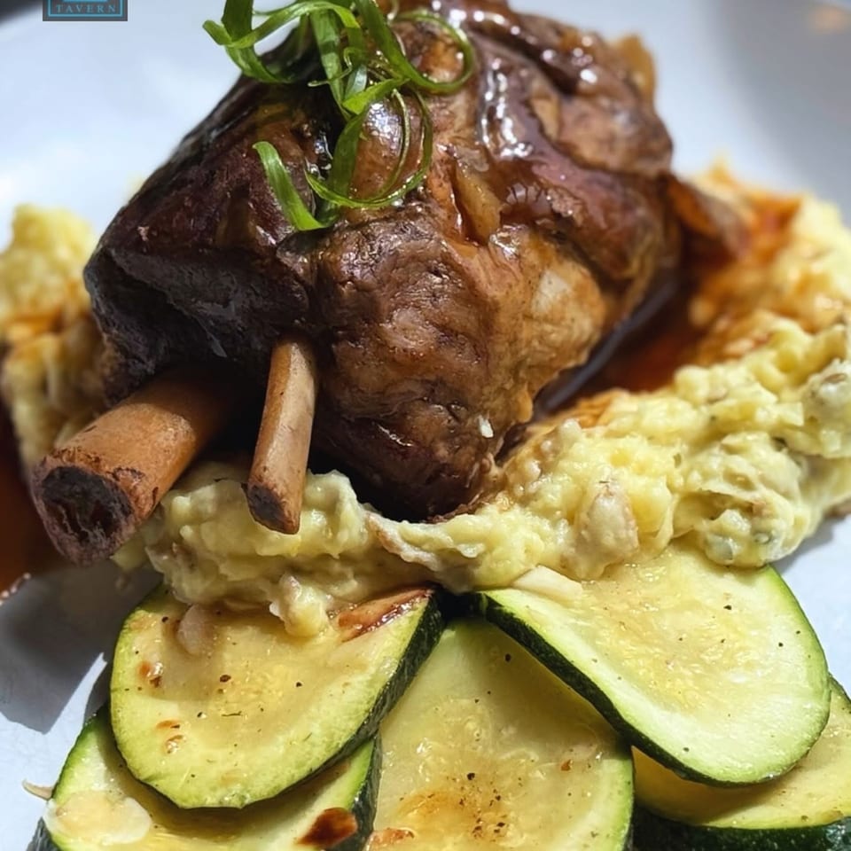 Braised lamb shank with mashed potatoes, gravy, and sliced zucchini on a white plate.