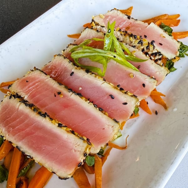Seared tuna slices with sesame seeds, green onions, and carrot strips on a white rectangular plate.