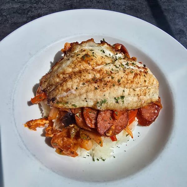 Grilled fish fillet over rice with sausage, shrimp, and vegetables in a white bowl.