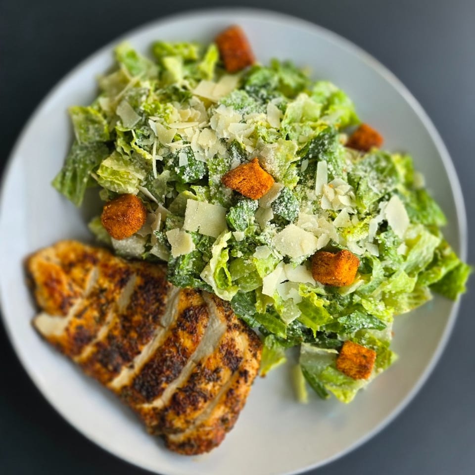 Grilled sliced chicken served with Caesar salad, Parmesan shavings, and golden croutons on a white plate.