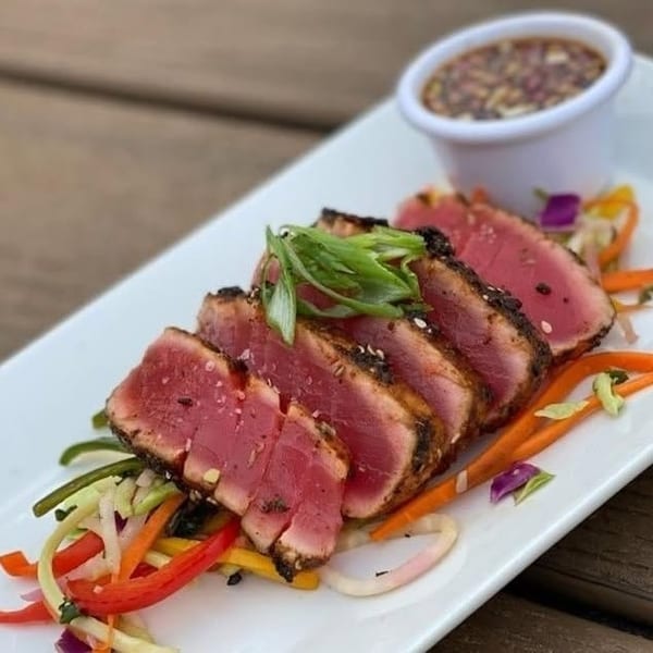 Seared tuna slices with sesame seeds, green onions, and colorful vegetables, served with dipping sauce.